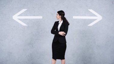 pensive businesswoman in suit looking at arrows on grey