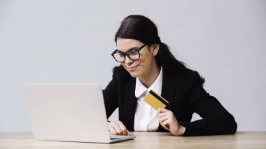 cheerful businesswoman holding credit card near laptop isolated on grey