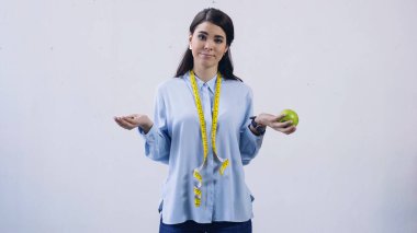 confused woman comparing vitamins and apple isolated on grey