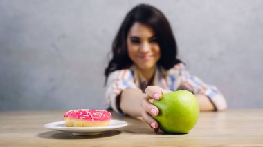 blurred woman taking green apple near doughnut on grey