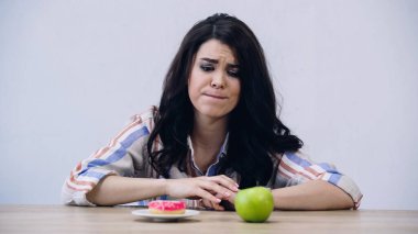 confused woman choosing between apple and doughnut isolated on grey