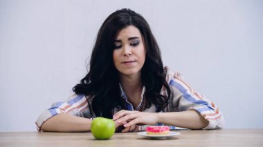 young woman choosing between apple and doughnut isolated on grey