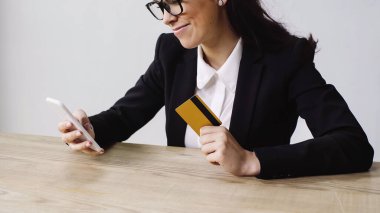 cheerful businesswoman doing online shopping while holding smartphone and credit card isolated on white 