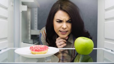 brunette woman choosing between apple and glazed doughnut in fridge 