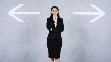pensive brunette businesswoman in suit standing near arrows on gray 