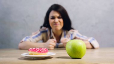 blurred woman looking at apple and doughnut on grey