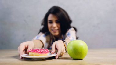 happy woman taking plate with sweet doughnut near apple on table on grey