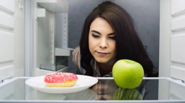 brunette woman choosing between fresh apple and doughnut in fridge