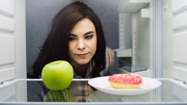 brunette woman choosing between green apple and doughnut in fridge 