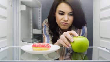 displeased woman taking green apple near doughnut in fridge
