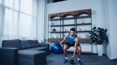 Mavi kolsuz tişörtlü sakallı genç sporcu evde kettlebell egzersizi yapıyor.