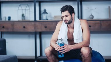 Havlulu yorgun sporcu spor topunun üzerinde oturuyor ve elinde spor şişesi tutuyor. 