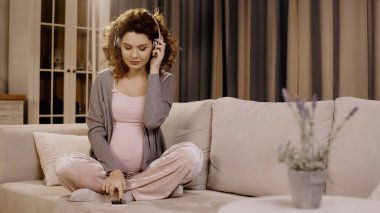 Pregnant woman in headphones using smartphone on couch at home 