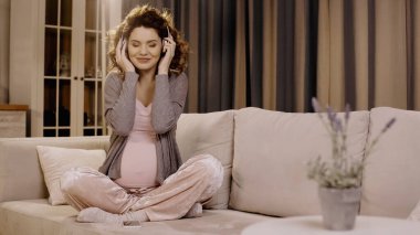 Cheerful pregnant woman in headphones sitting near smartphone on couch 