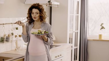 Mutfakta taze salata ve çatal tutan neşeli hamile kadın. 