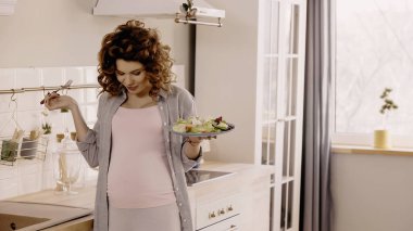 Mutfakta karnına bakarken elinde çatal ve salata tutan kıvırcık hamile kadın. 