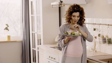 Genç hamile kadın mutfakta taze salata ve çatal tutuyor. 