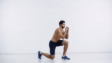 Beyaz tuğla duvarın yanında spor yaparken şort ve spor ayakkabısı giyen sakallı bir sporcu.