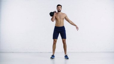 Üstsüz ve sportif bir adam beyaz tuğla duvarın yanında ağır bir kettlebell ile egzersiz yapıyor. 