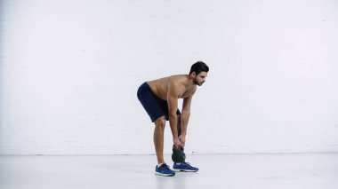 Sakallı ve sportif bir adam beyaz tuğla duvarın yanında ağır bir kettlebell ile egzersiz yapıyor. 