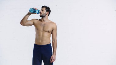 Mavi şortlu kaslı sporcu beyaz tuğla duvarın yanındaki spor şişesinden su içiyor. 