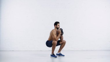 Beyaz tuğla duvarın yanında ağır kettlebell ile spor yapan üstsüz bir sporcu.