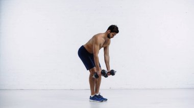 Beyaz tuğla duvarın yanında ağır dambılları olan şortlu üstsüz bir sporcu.