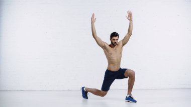 Tam boy kaslı sporcu beyaz tuğla duvarın yanında ellerini kaldırarak hamle yapıyor. 