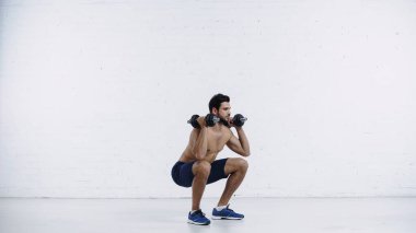 Uzun boylu, şortlu, beyaz tuğlalı duvara dambıllarıyla çömelmiş bir adam. 