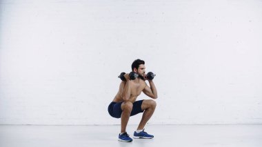 full length of sportive man in shorts squatting with dumbbells against white brick wall  clipart