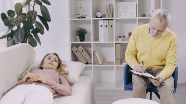 Orta yaşlı psikoterapistin yanındaki kanepede yatan üzgün kadın panoya yazı yazıyor. 