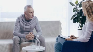 Orta yaşlı, spor giyimli, elleri sımsıkı bağlı bir adam psikoterapistin yanındaki koltukta oturuyor. 