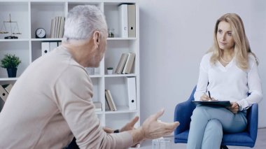 Sarışın psikolog, muayene sırasında konuşan olgun bir adama bakıyor. 