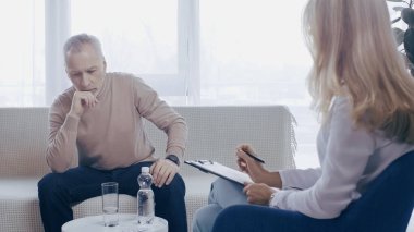 Sarışın psikolog, danışma odasında düşünceli bir adamın yanında kalem ve not defteri tutuyor.