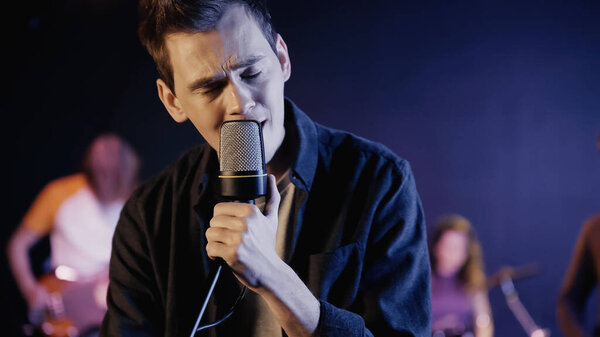 blurred young singer with closed eyes singing near music band on stage 