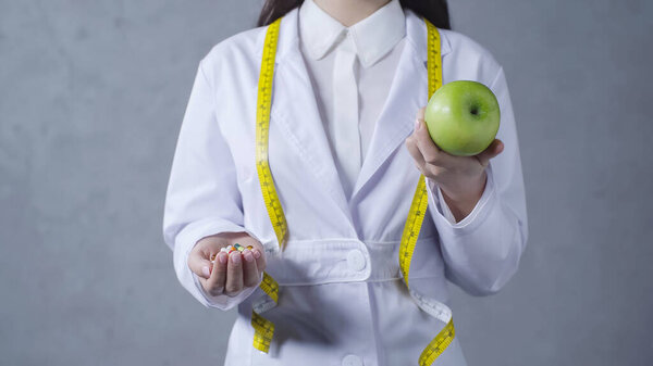 cropped view of nutritionist comparing apple and vitamins on grey