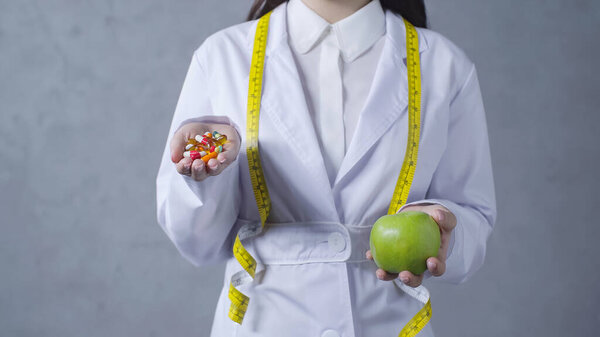 partial view of nutritionist comparing apple and vitamins on grey 