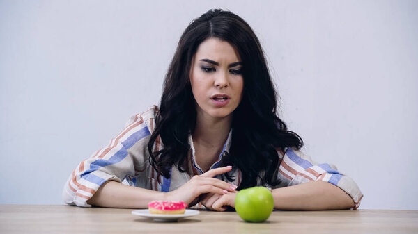 brunette woman choosing between apple and doughnut isolated on grey