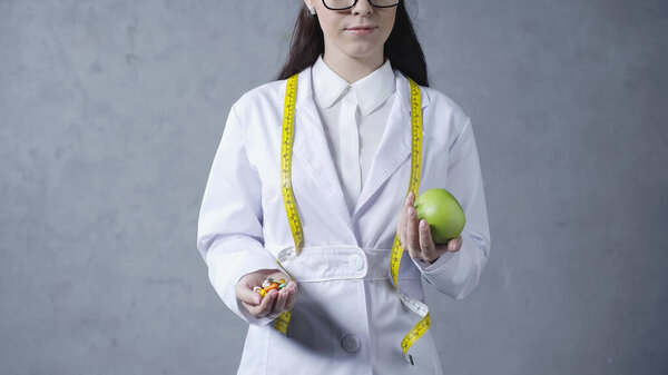 partial view of brunette nutritionist in white coat comparing apple and vitamins on grey 