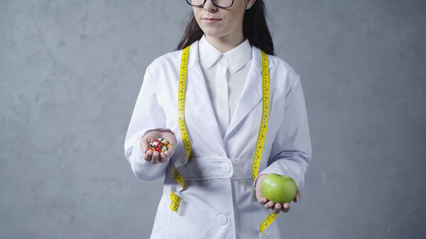 partial view of nutritionist in white coat comparing apple and vitamins on grey 