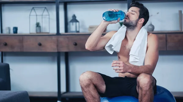 exhausted sportsman with towel sitting on fitness ball and holding sports bottle while drinking water