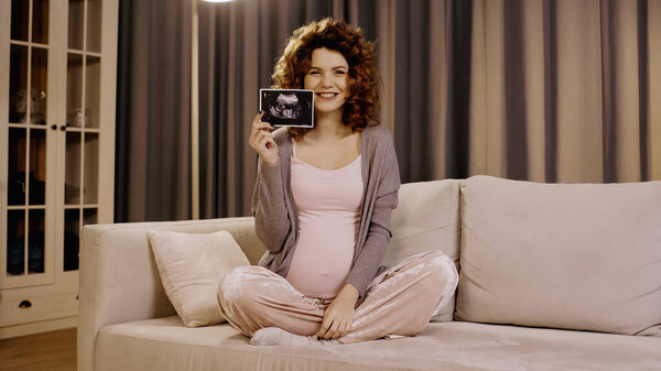Smiling pregnant woman holding ultrasound scan of baby on couch 
