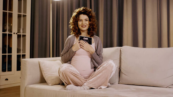 Smiling pregnant woman holding ultrasound scan of baby on couch in living room 