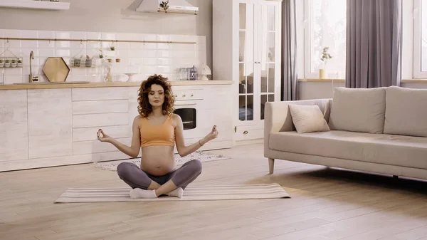 坐在瑜伽上的卷曲孕妇在厨房的垫子上摆姿势 — 图库照片