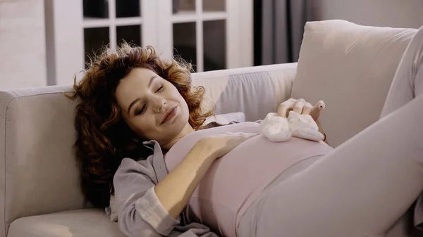 抱着婴儿靴靠近肚子躺在沙发上的快乐孕妇 — 图库照片
