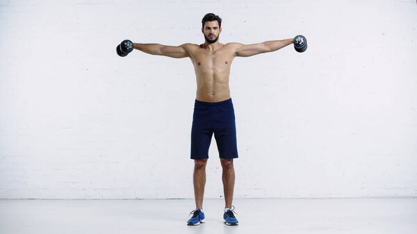 full length of shirtless sportsman with outstretched hands holding heavy dumbbells near white brick wall 