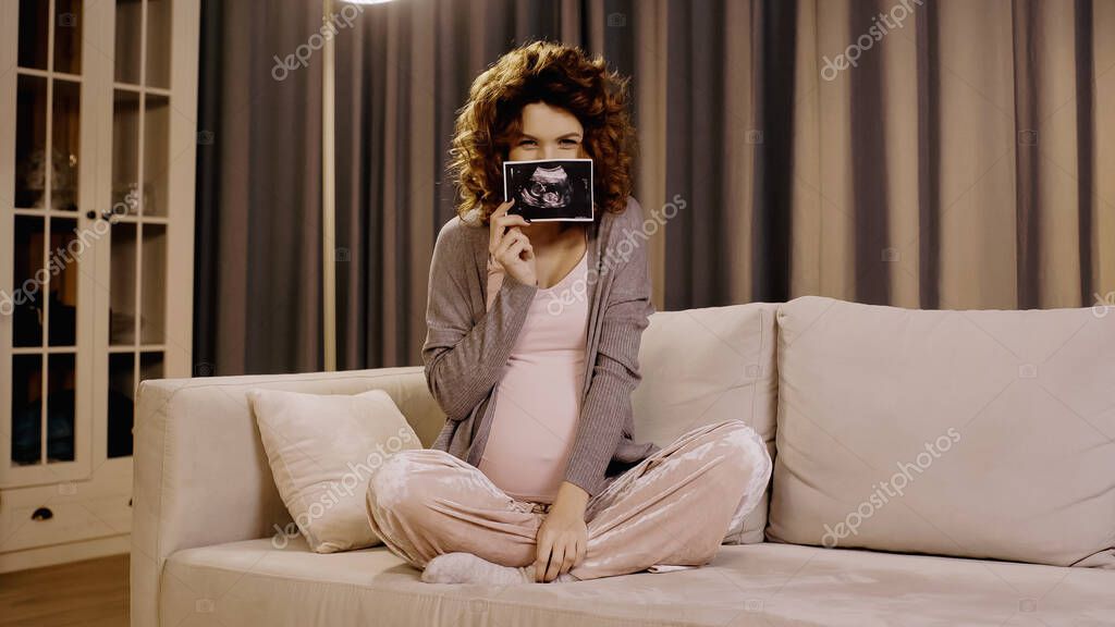 Pregnant woman covering face with ultrasound scan of baby at home
