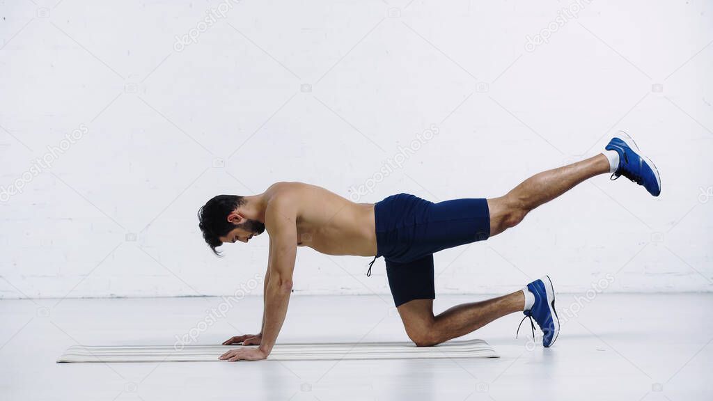 side view of shirtless sportsman doing knee plank on fitness mat near white brick wall 