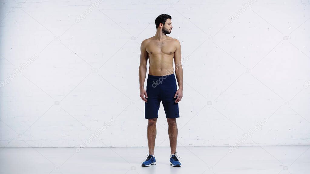 full length of sportsman in shorts standing and doing neck exercise near white brick wall