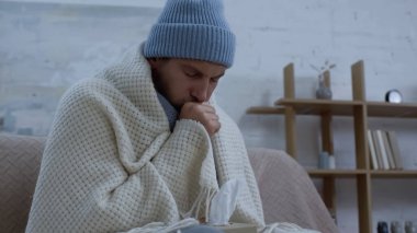 Sıcak şapkalı ve battaniyeli hasta adam evdeki kanepede öksürüyor.